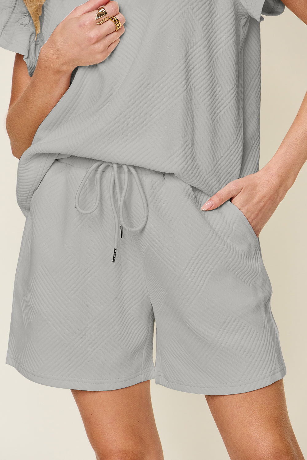 Against a plain background, a woman with blonde hair poses confidently with one hand in her pocket, highlighting the chic style of her Double Take Full Size Texture Flounce Sleeve Top and Drawstring Shorts Set.