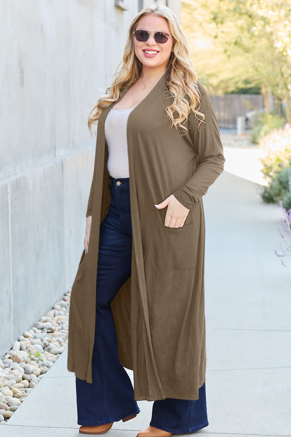 A woman stands against a concrete wall, wearing the Basic Bae Full Size Open Front Long Sleeve Cover Up made from rayon spandex, a white top, blue jeans in her perfect size, and brown shoes with her hands in her pockets. Machine wash cold for best results.