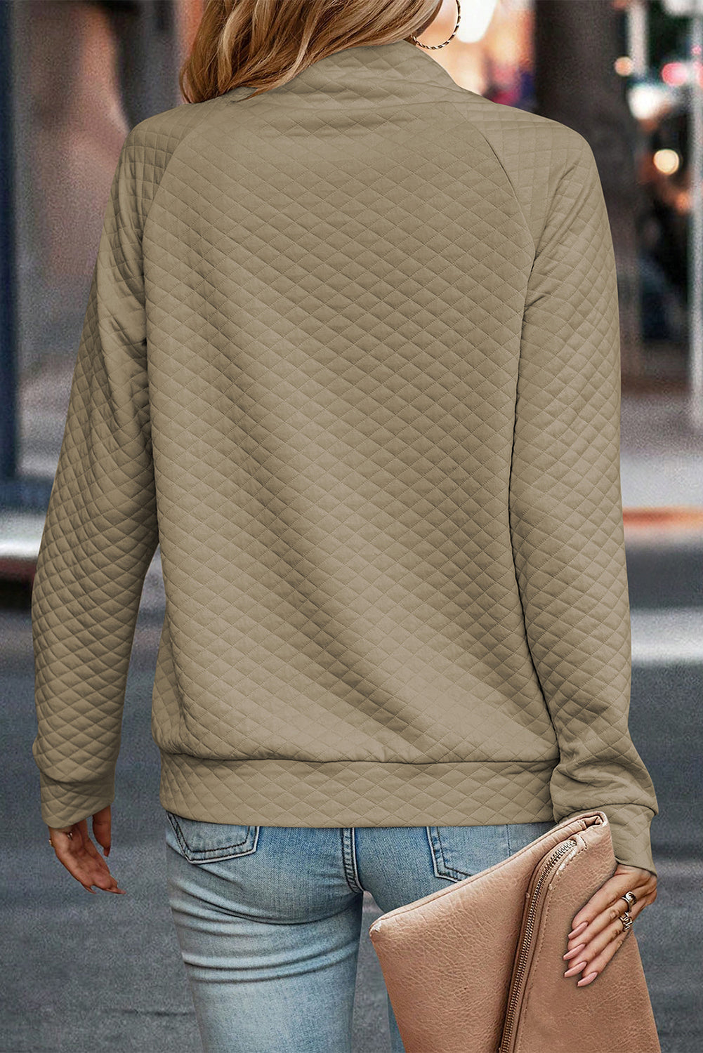 A person wearing an Apricot Quilted Buttoned Neckline Stand Neck Pullover Sweatshirt with light blue jeans holds a beige clutch while walking away on a city sidewalk.