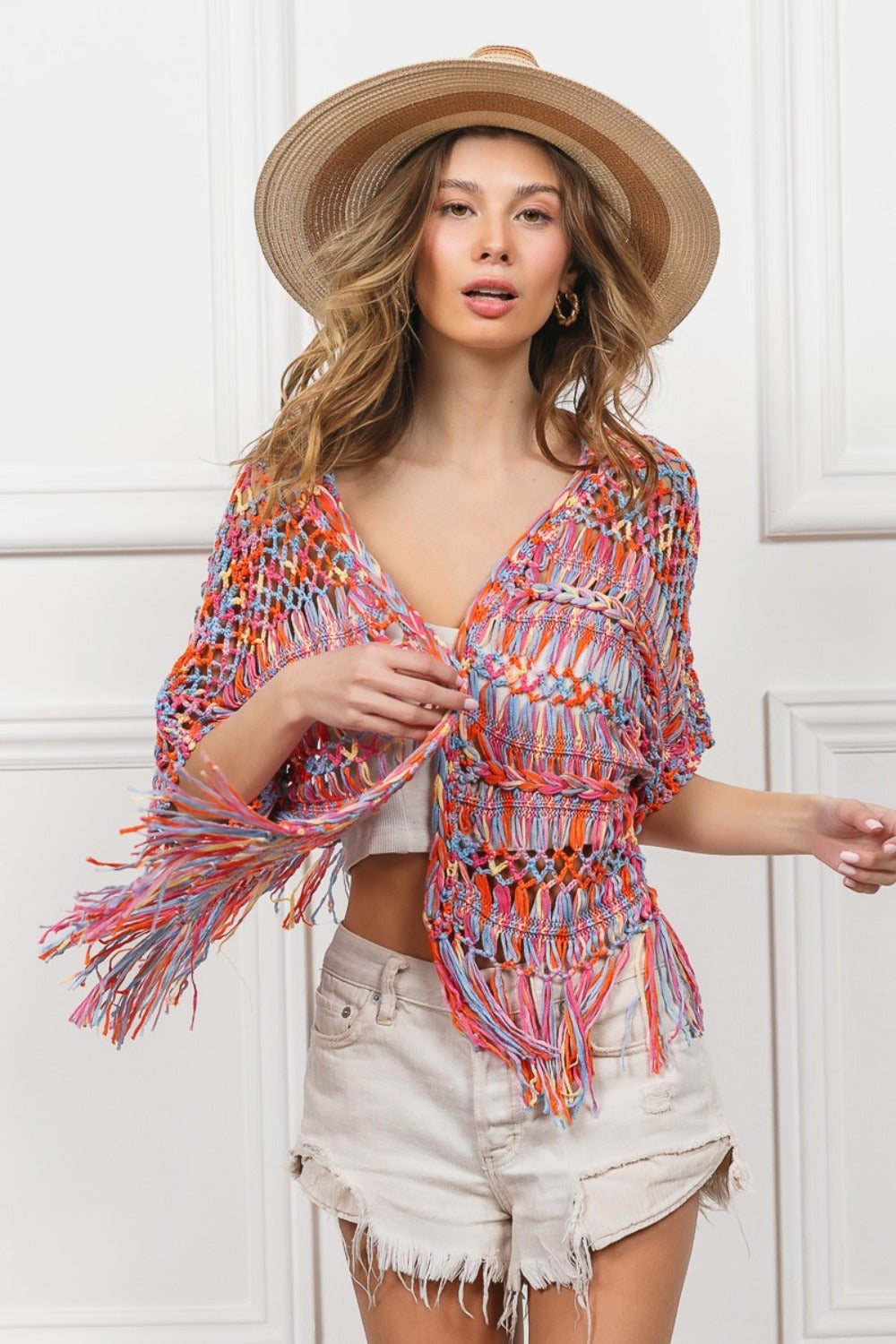 A woman wearing a BiBi Open Front Fringed Crop Knit Cardigan, a white bandeau, distressed white shorts, and a wide-brimmed straw hat stands against a white paneled background, smiling with one hand on her hat.