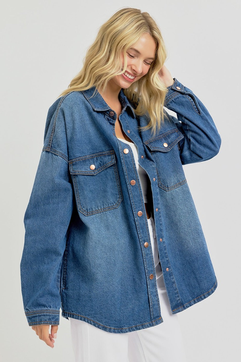 A person with long blonde hair, wearing the RISEN Full Size Snap Down Long Sleeve Denim Shacket over a white crop top and white pants, stands against a plain background.