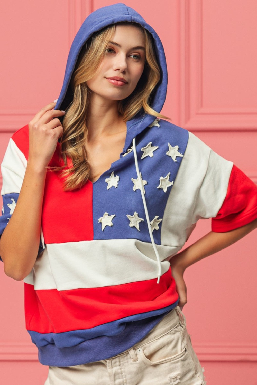 Woman wearing a BiBi American Flag Theme Hoodie, standing against a pink background.