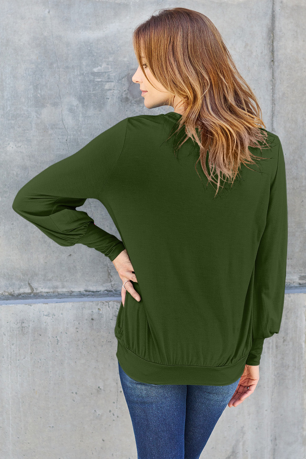 A woman with brown hair is standing against a concrete wall, wearing the Basic Bae Full Size V-Neck Lantern Sleeve Top in teal and blue basic-style jeans, smiling.