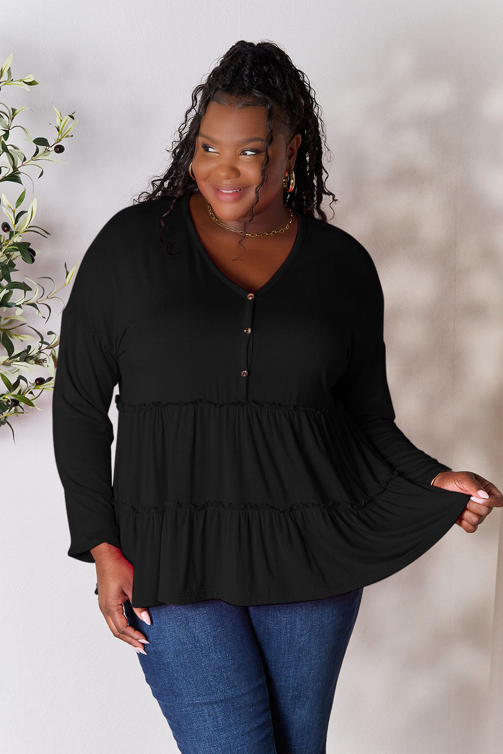 Person stands indoors near a plant, wearing the Double Take Half Button Long Sleeve Ruffle Hem Blouse in gray and blue jeans, smiling while looking to the side. The slightly stretchy fabric of the blouse adds comfort while its buttoned design offers a touch of elegance.