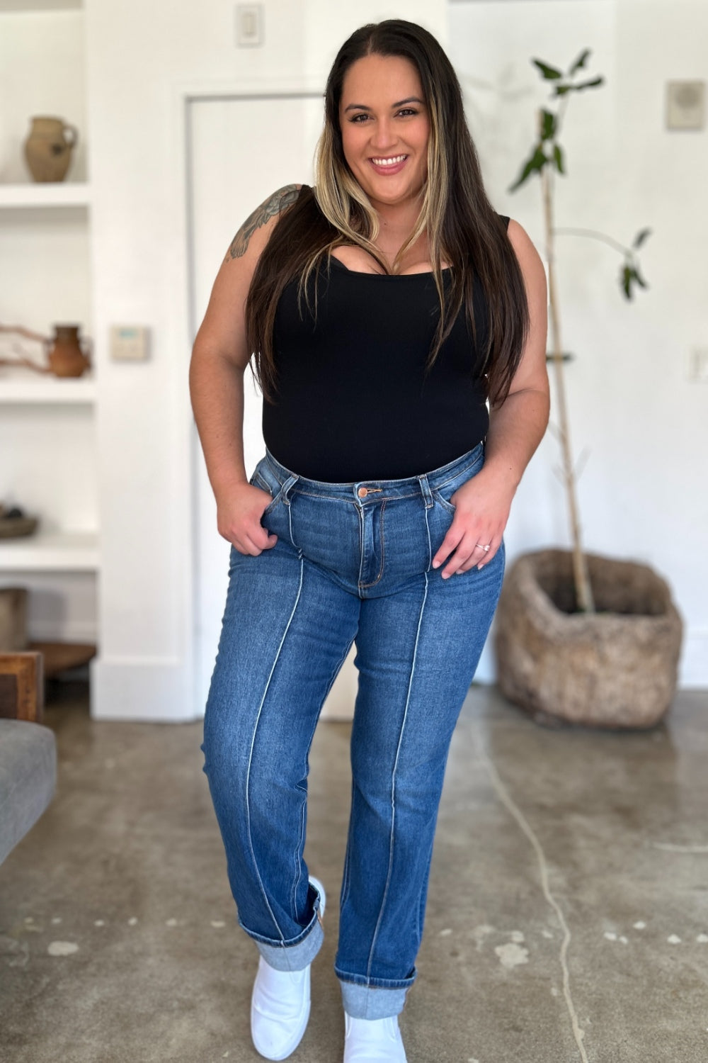 Two women stand side by side indoors, both wearing black tank tops, Judy Blue Full Size High Waist Front Seam Detail Straight Jeans, and white shoes. They are smiling and posing for the camera. A plant and shelves with decorations are in the background.