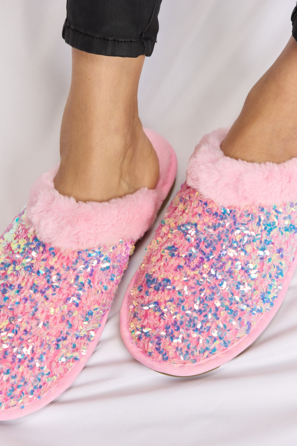 Person wearing cozy and luxurious Forever Link Sequin Plush Round Toe Slippers with fuzzy trim and black pants, sitting on a white surface.