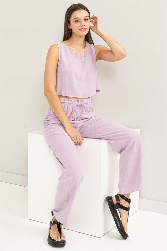 A person is seated on the floor, dressed in a soft lavender D-Linen Blended Top and Pants Set, complemented by chic black sandals.