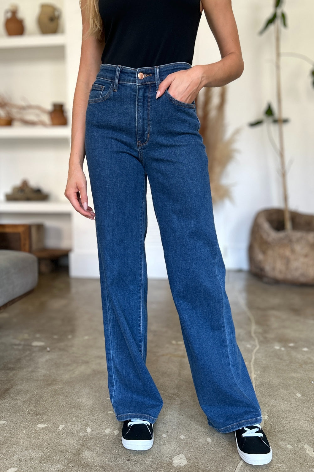 A person wearing Judy Blue Full Size High Rise Straight Jeans, a black top, and white shoes stands gracefully on a polished concrete floor.