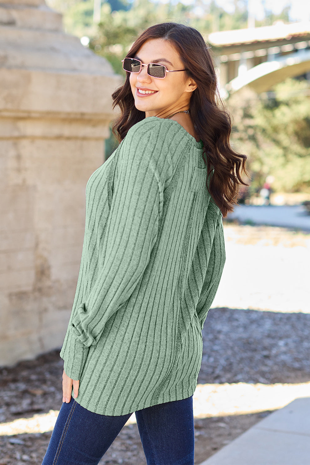 A person with long dark hair and sunglasses stands outdoors, wearing the Basic Bae Full Size Ribbed Round Neck Long Sleeve Knit Top in blue and a pair of jeans. The basic style outfit blends seamlessly with the background of trees and a bridge.