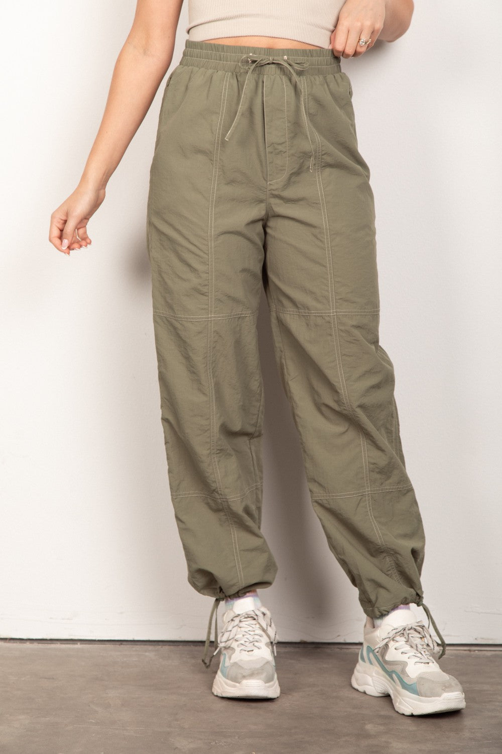 A person wearing VERY J Drawstring Woven Parachute Joggers in olive green and white sneakers stands against a plain background, showcasing the seamless blend of style and comfort with intricate woven fabric details.