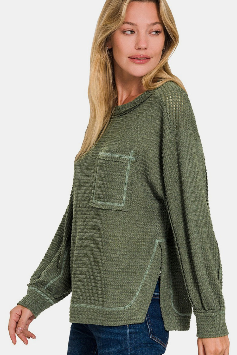 A person wearing the Zenana Exposed Seam Side Slit Long Sleeve Top in textured green, paired with blue jeans, stands against a plain background.