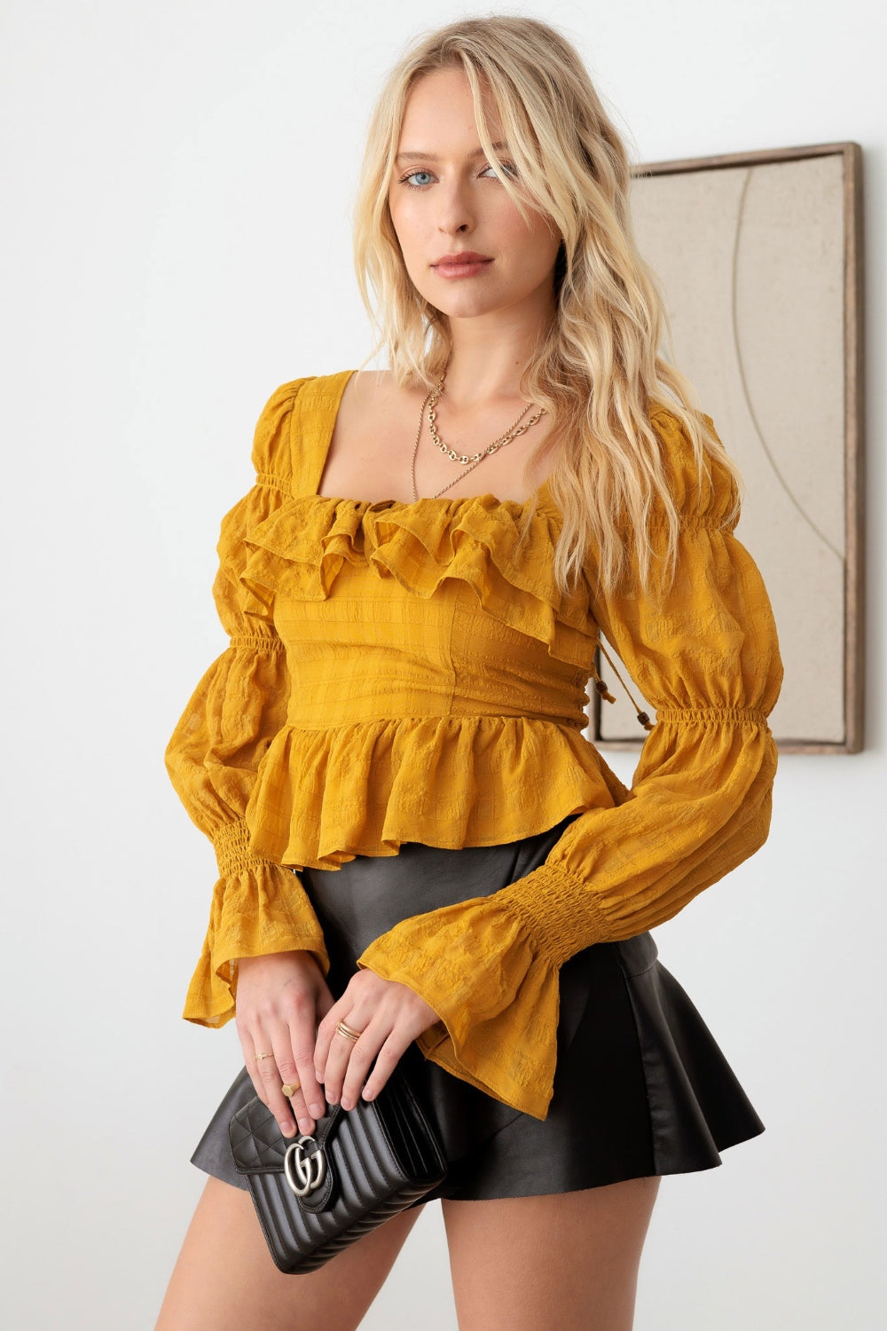 A woman with long blonde hair is wearing the Mustard Seed Boho Shirred Smocked Peplum Long Sleeve Top, a black skirt, and holding a black purse. She is standing in front of a white wall adorned with framed art.