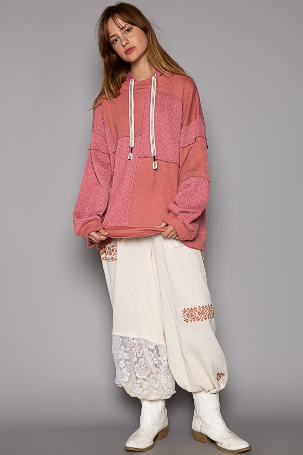 A person wearing the POL Exposed Seam Hooded Knit Top, featuring a pink patchwork design with balloon sleeves and white drawstrings, paired with relaxed-fit white pants, poses against a gray background.