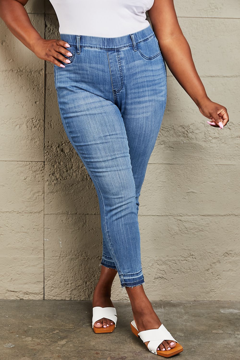 Person wearing Judy Blue Janavie Full Size High Waisted Pull On Skinny Jeans with release hem detailing and white sandals stands against a textured wall.