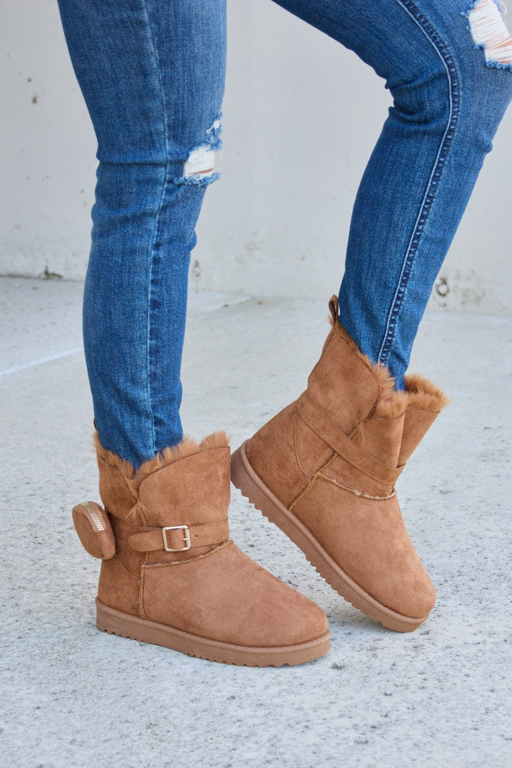 Person wearing ripped blue jeans and Forever Link Plush Thermal Flat Boots, tan in color, fur-lined with side buckles, standing on a concrete surface. These stylish boots promise cozy insulation for the feet.