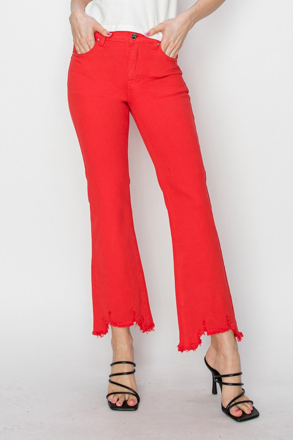 A person is wearing the RISEN Raw Hem Bootcut Jeans with Pockets in bright red, paired with a white top and black strappy high-heeled sandals. The image focuses on the stylish and functional lower half of the body.