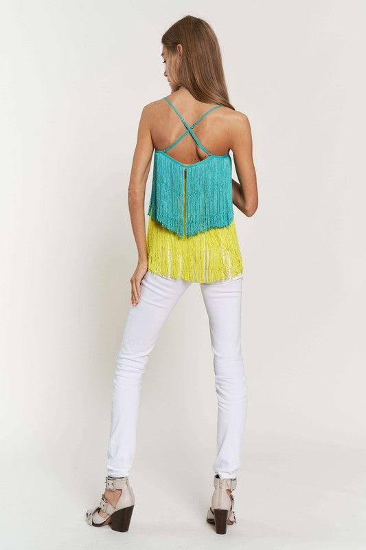 A woman with long brown hair is wearing a Fringe Overlay Cross Straps Party Cami Top featuring blue and yellow layers, paired with white pants, standing against a plain background.