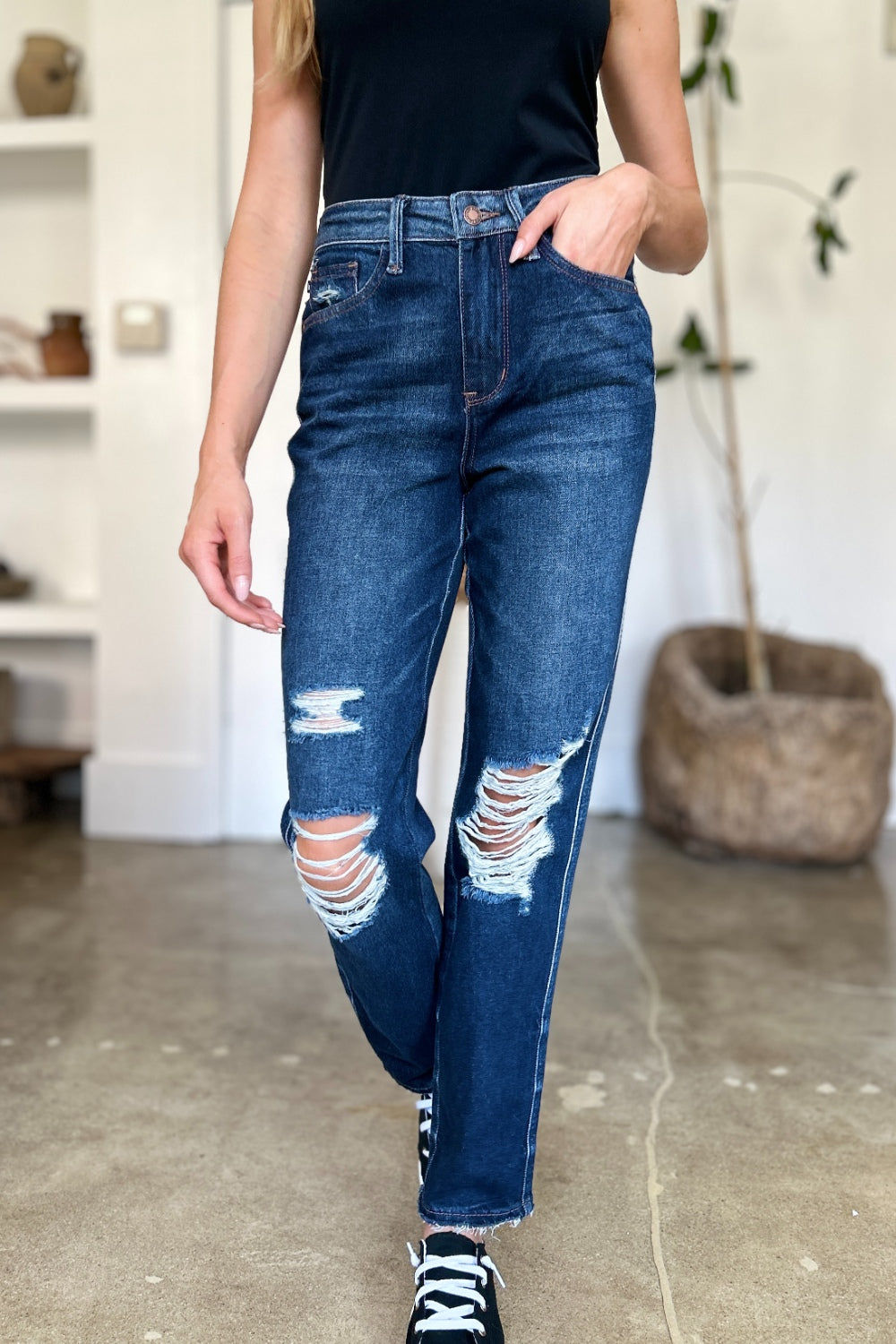 A woman in a black sleeveless top and Judy Blue Full Size High Waist Rigid Magic Heavy Destroy Straight Jeans stands indoors with her hands in her pockets. The background features a plant and shelves adorned with decor items.