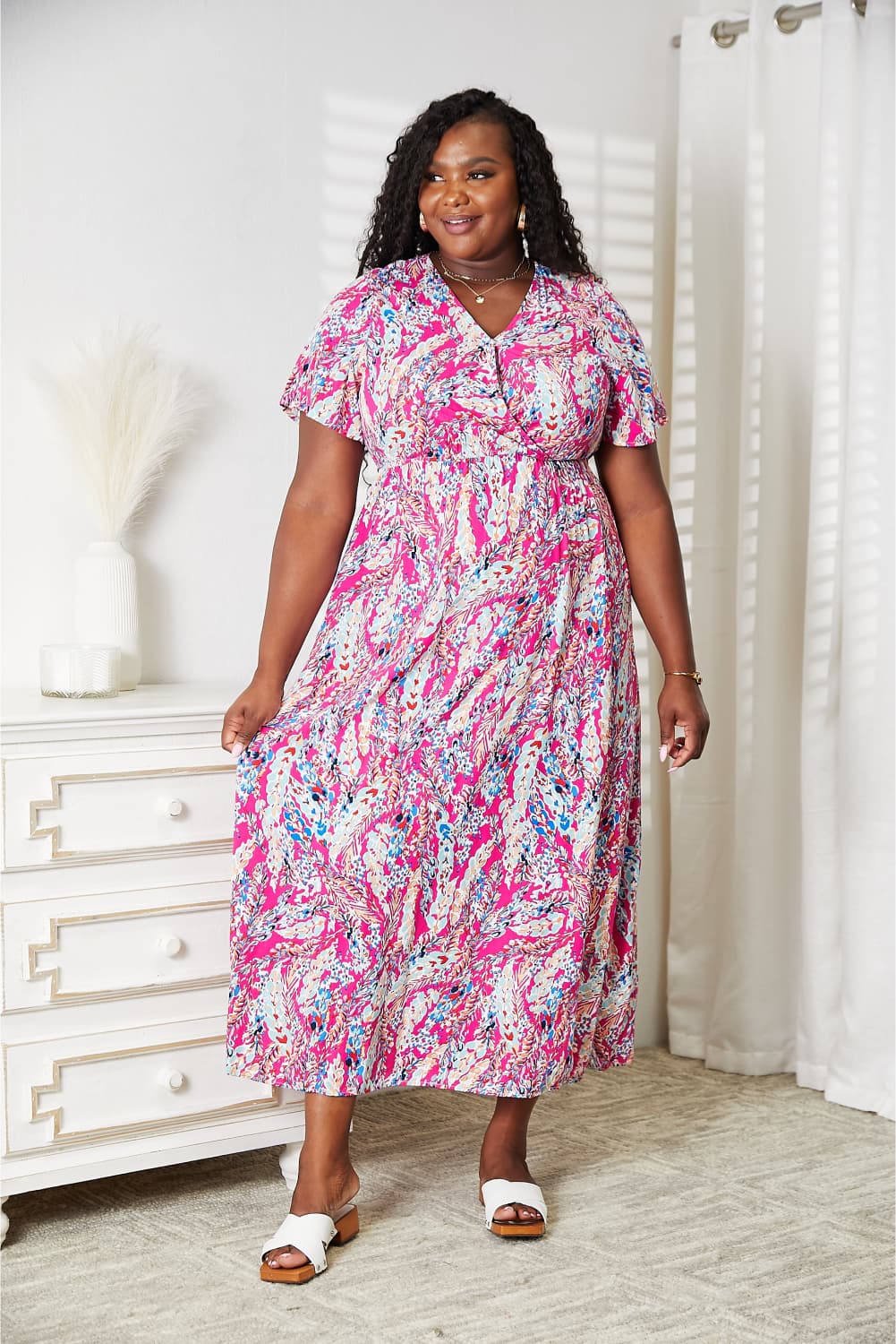 A woman stands in a room wearing the Double Take Multicolored V-Neck Maxi Dress, paired with white sandals. A dresser and curtain create a charming backdrop.