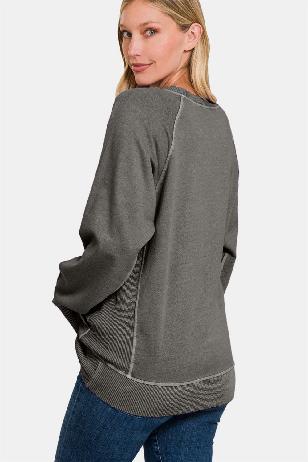 A person stands wearing a Zenana Round Neck Long Sleeve Sweatshirt, featuring a loose fit, dark grey color with front pockets and light stitching, paired with blue jeans. The 100% cotton fabric ensures comfort and durability.