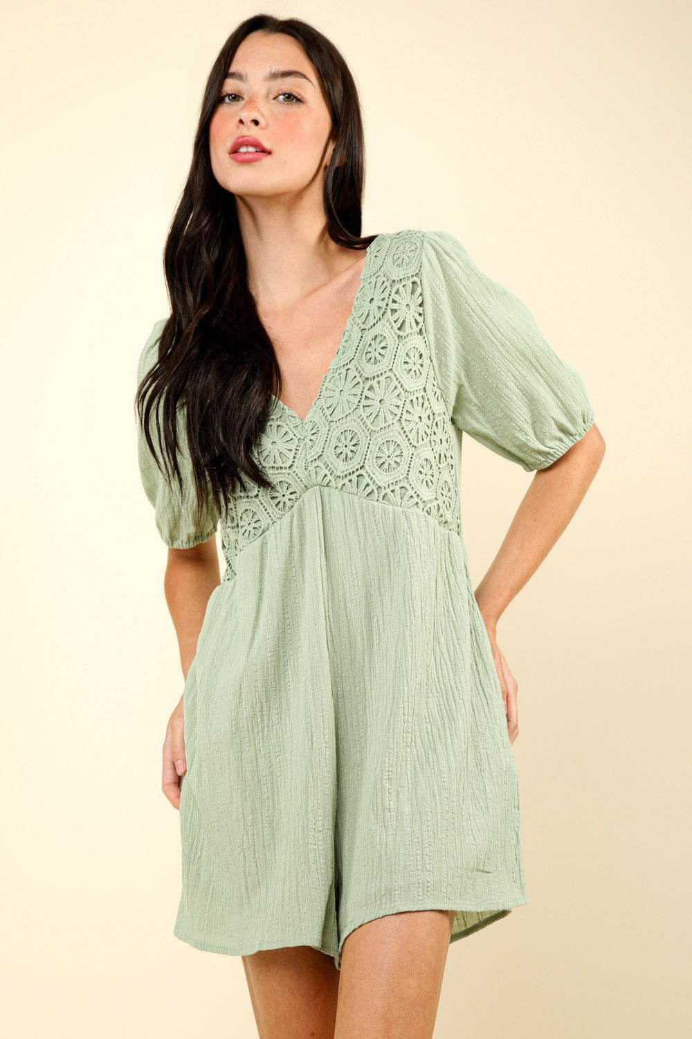 A person with long dark hair wearing a VERY J Lace Detail Puff Sleeve Romper with Pockets in light green, featuring a patterned bodice, stands against a plain beige background.