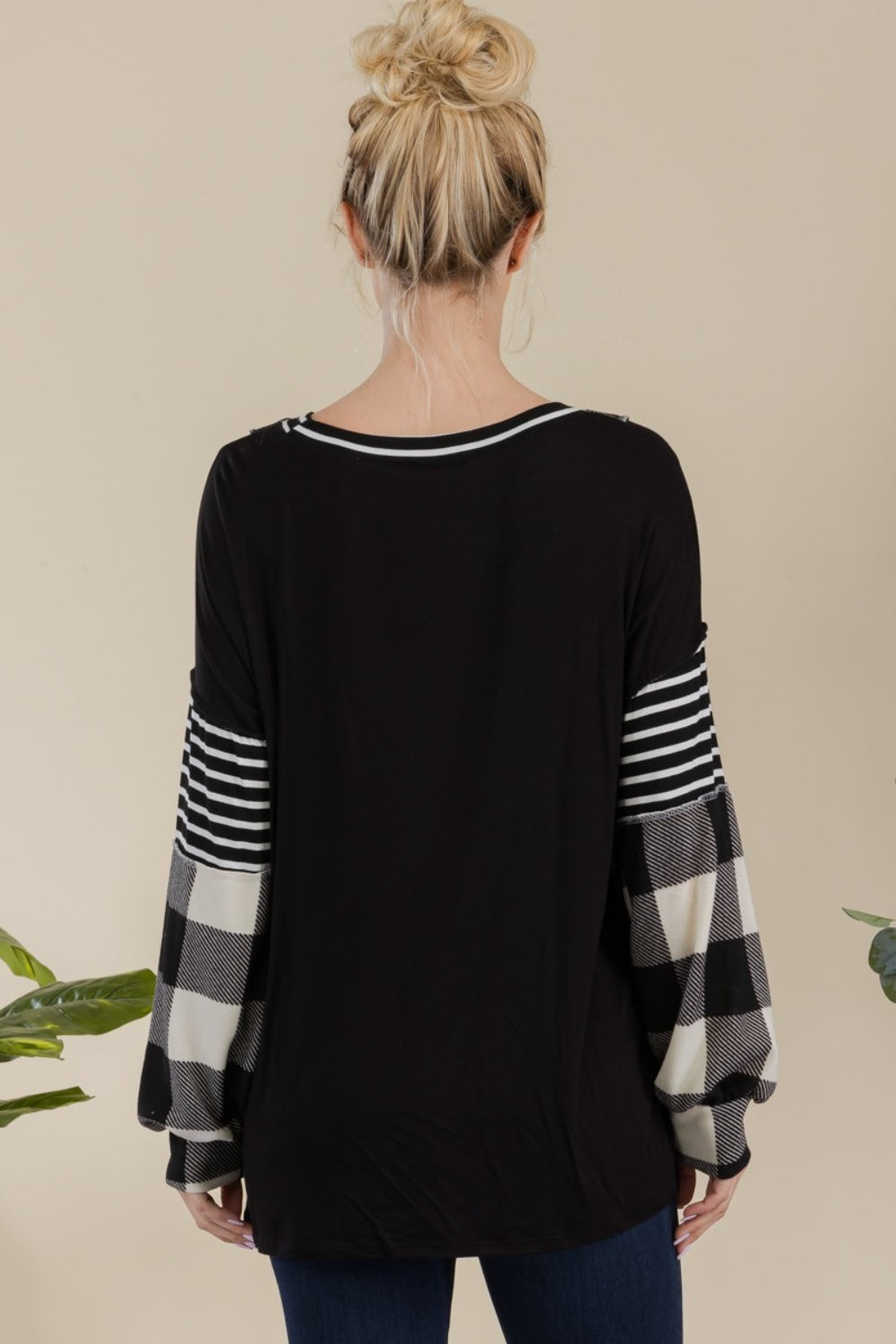 A person wearing a Celeste Full Size Color Block Exposed Seam T-Shirt, featuring black and white plaid with reverse stitching and long sleeves, poses while smiling in front of a neutral background with a plant.