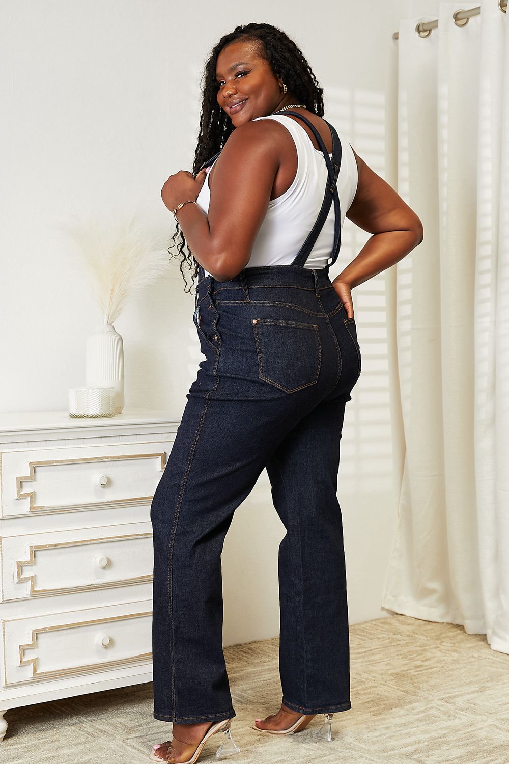 A person stands indoors wearing Judy Blue Full Size High Waist Classic Denim Overalls and a white tank top, with one hand in the pocket and the other resting on a white chest of drawers, next to a vase of dried pampas grass.