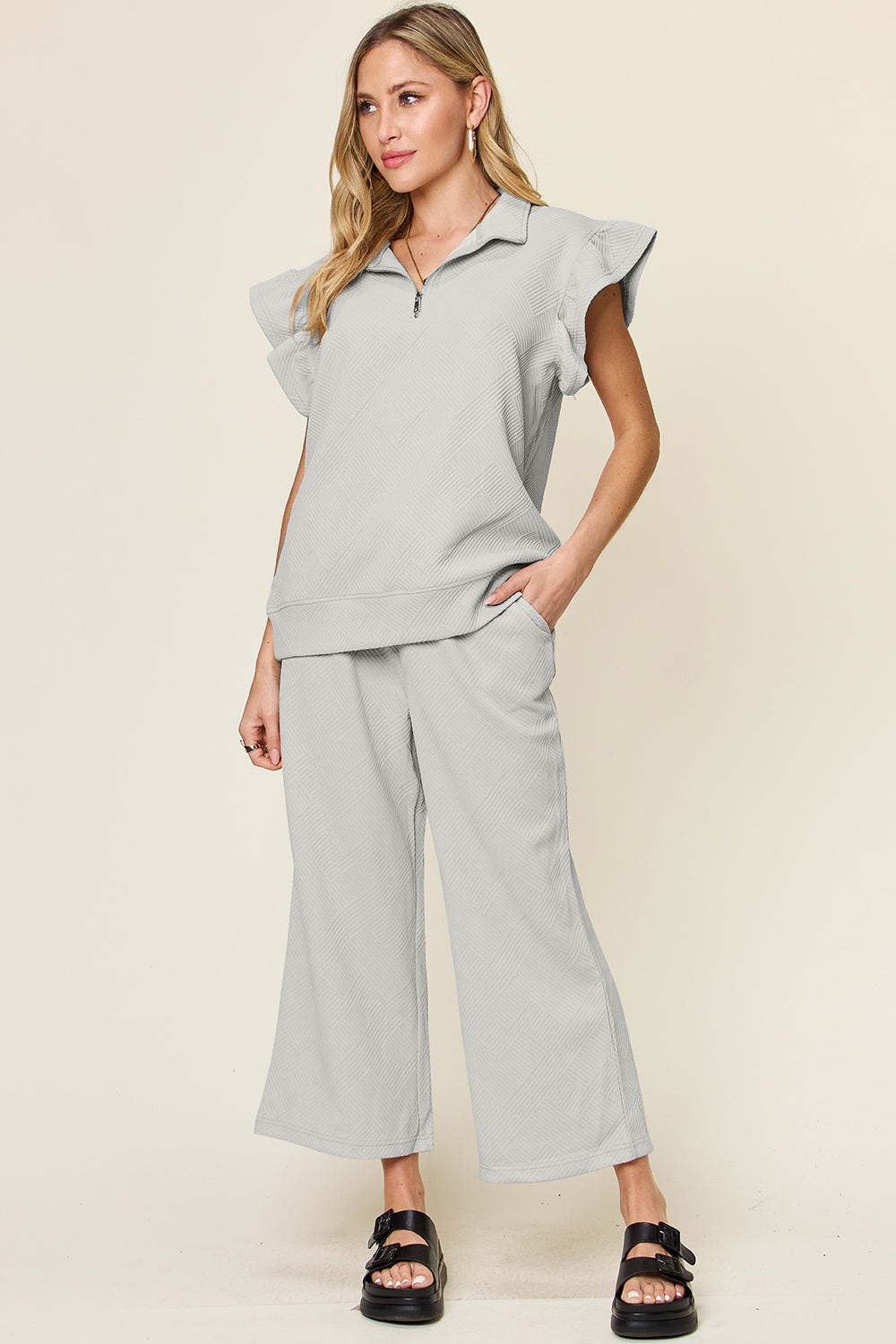 A woman stands wearing the Double Take Texture Ruffle Short Sleeve Top and Drawstring Wide Leg Pants Set in a slightly stretchy, light gray fabric with zipper detail. The two-piece outfit is complemented by black sandals, and she has one hand in her pocket. Machine wash cold for easy care.