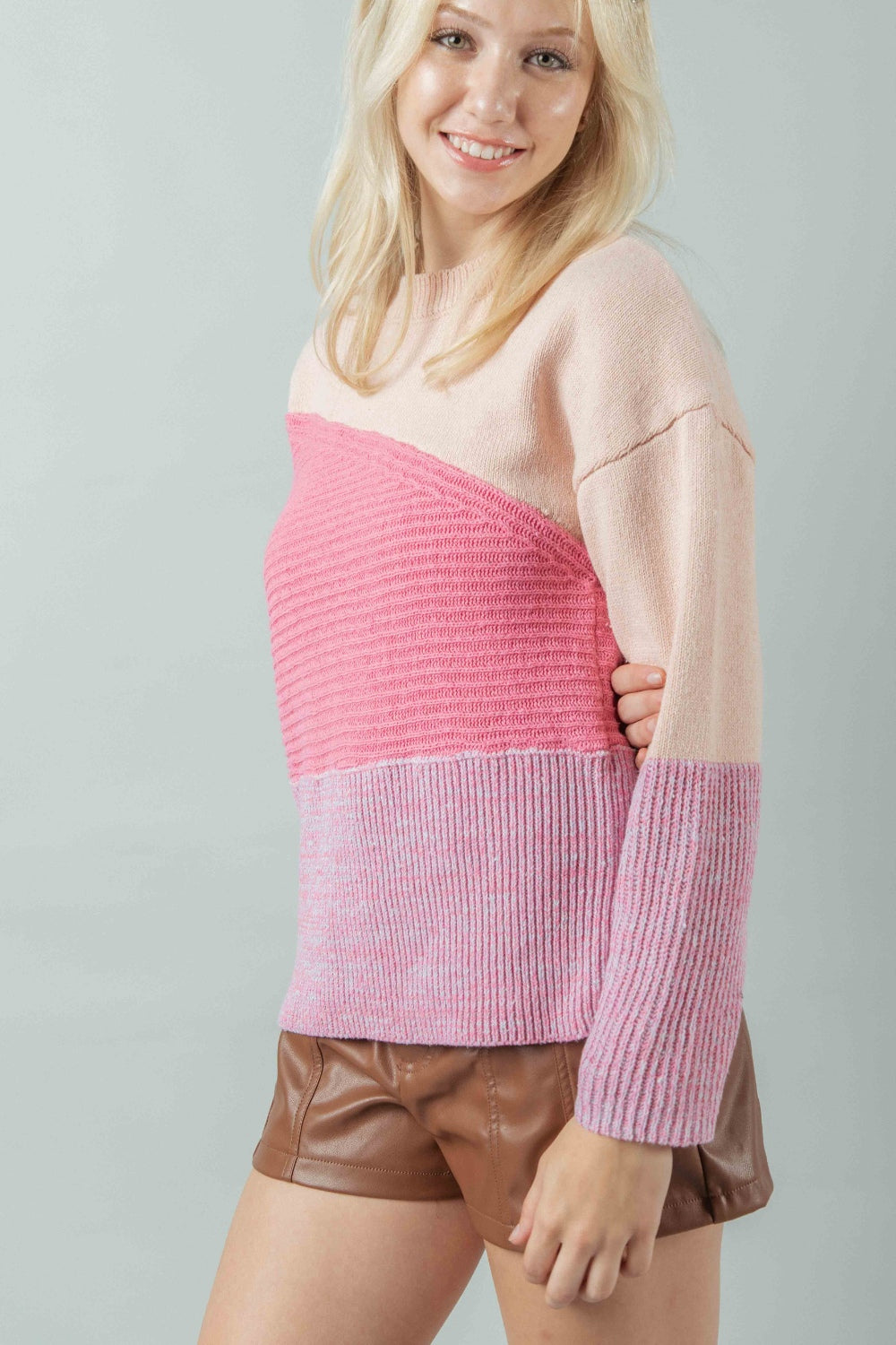 A woman stands against a plain background, elegantly dressed in a pink VERY J Color Block Long Sleeve Sweater and brown shorts, showcasing a chic blend of style and comfort.