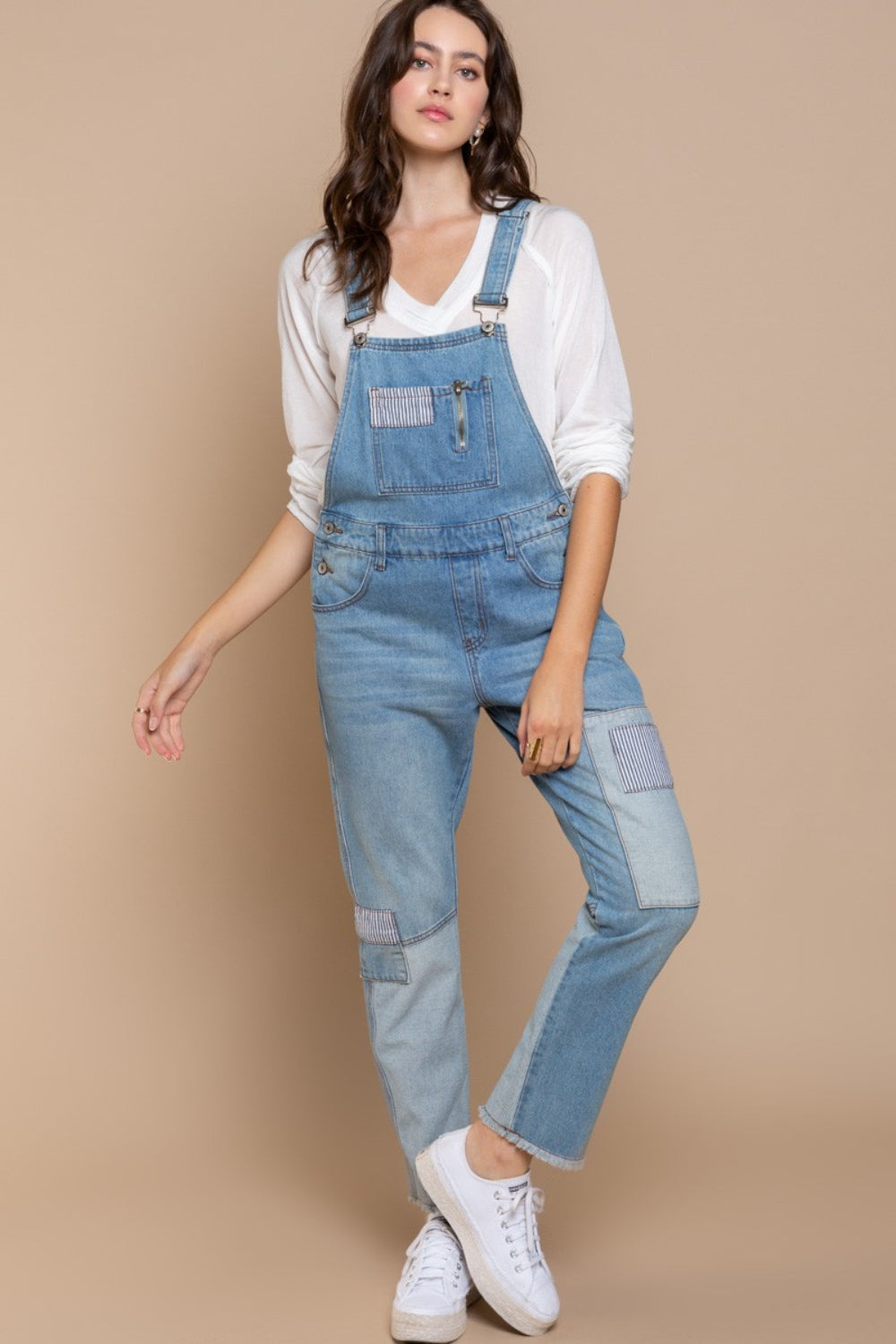 A woman kneels on one knee against a beige background, wearing a beige hat and white long-sleeve shirt, paired with POL Front Chest Zipper Slim Leg Denim Overalls featuring patches.