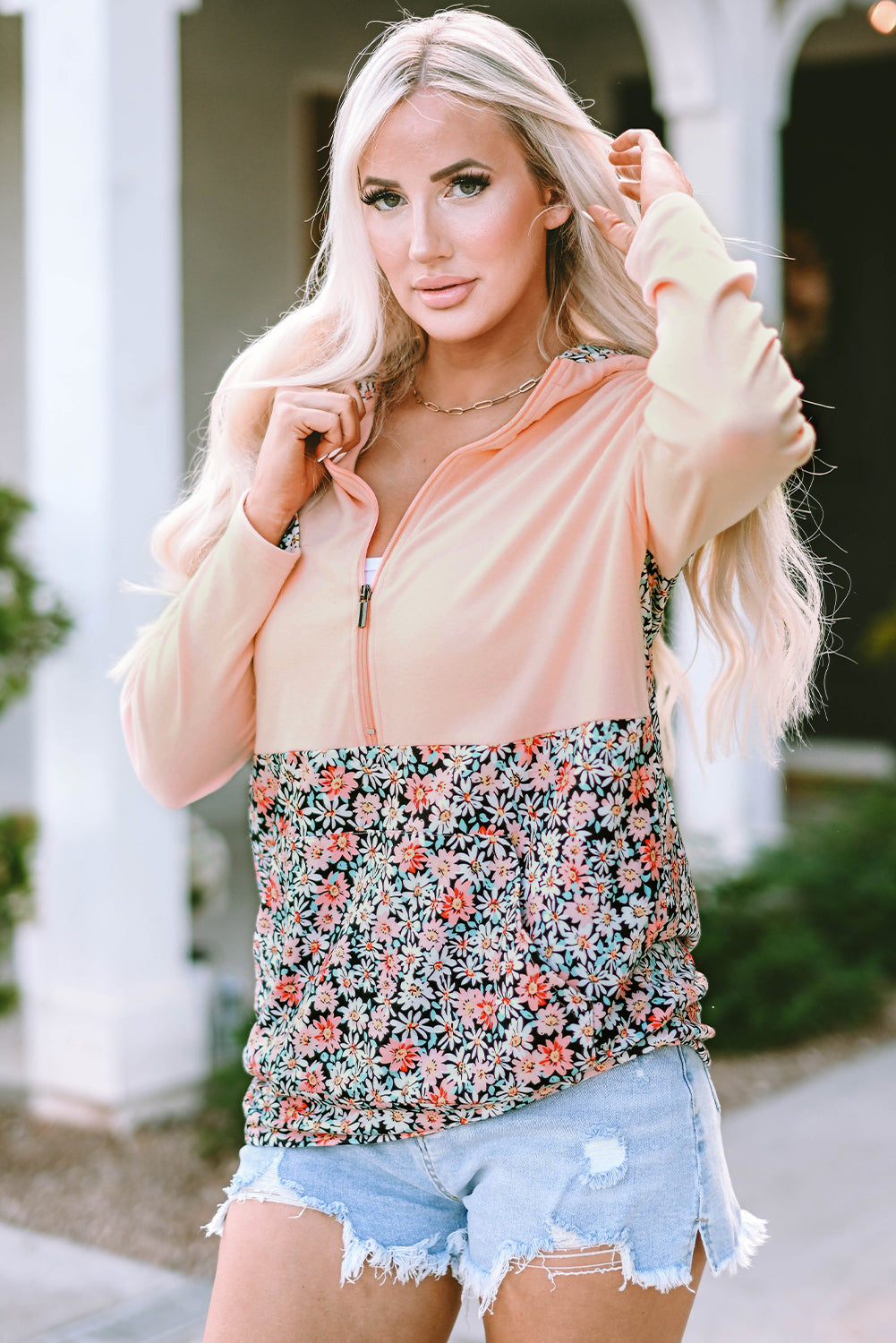 A person with long blonde hair wears a Pink Floral Patch Half Zip Kangaroo Pocket Hoodie and denim shorts, standing outdoors with their back facing the camera.