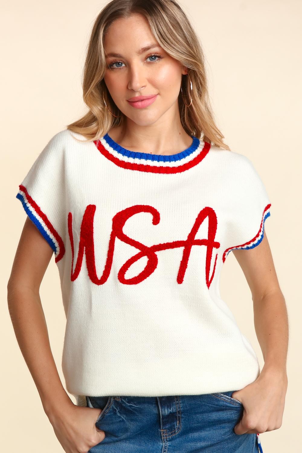 A person wearing the Haptics Letter Embroidery Slit Knit Top, a sleeveless white knit top with "USA" in red letter embroidery, accented with blue and red trim, and blue denim shorts, smiling at the camera.