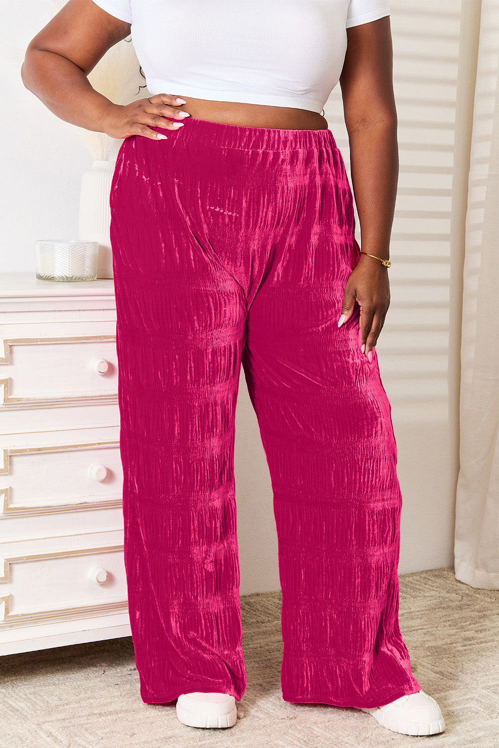 A person sporting the Double Take Full Size High Waist Tiered Shirring Velvet Wide Leg Pants in magenta, featuring an elasticized waistband, stands with hands in pockets in a room filled with white furnishings and natural lighting. The comfortable fit of the pants enhances the relaxed ambiance of the space.