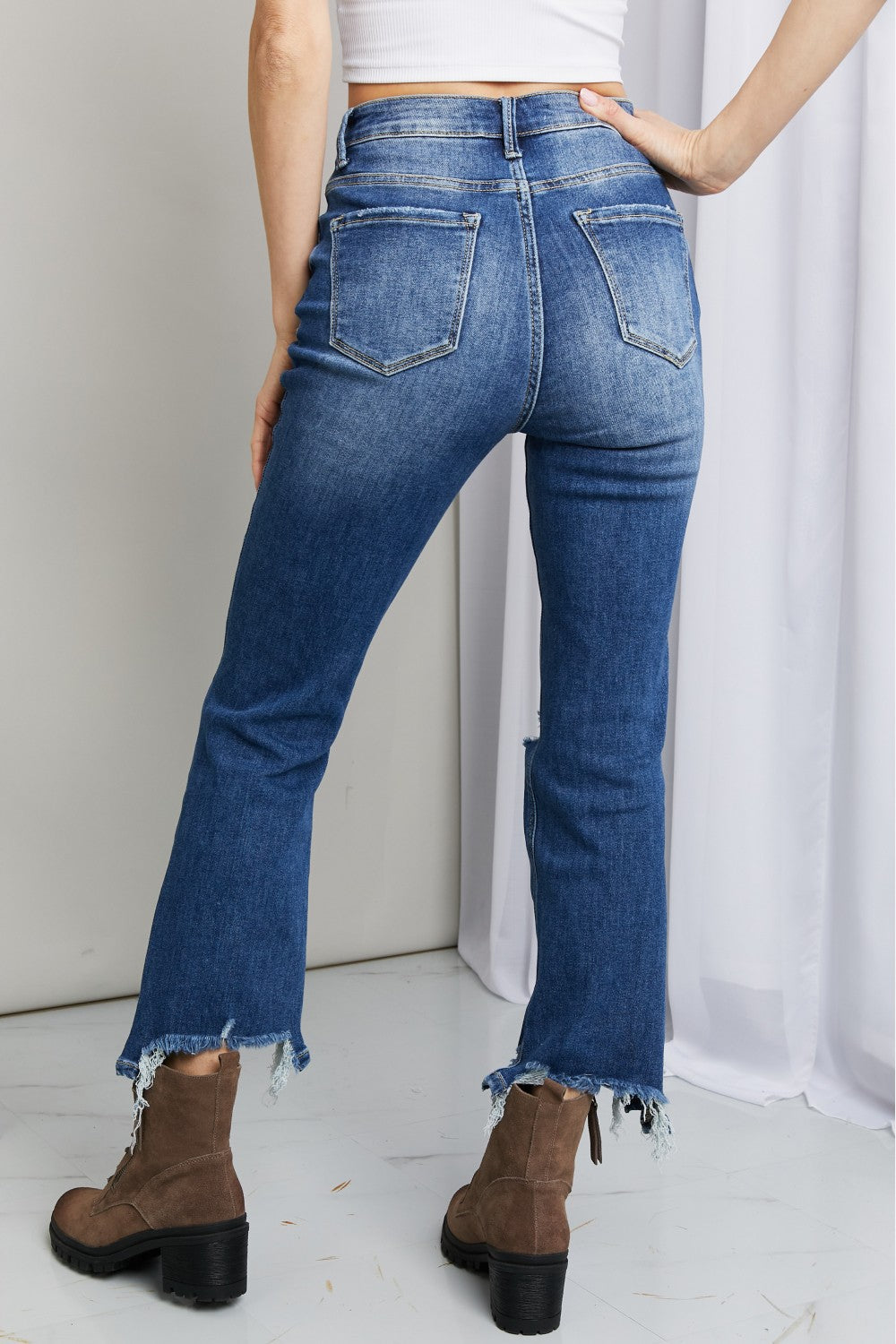 A person wearing RISEN Frayed Hem Distressed Cropped Jeans and brown ankle boots with a block heel stands in front of a white curtain next to a gold shelving unit.