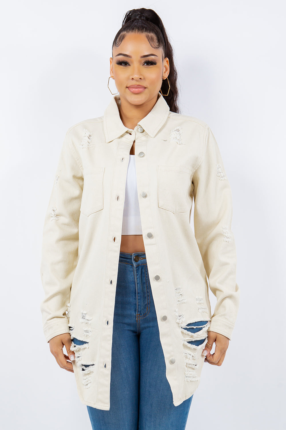 A woman wearing an American Bazi Distressed Button Up Long Sleeve Denim Jacket in beige, paired with a button-up white crop top and blue jeans, stands against a plain background.