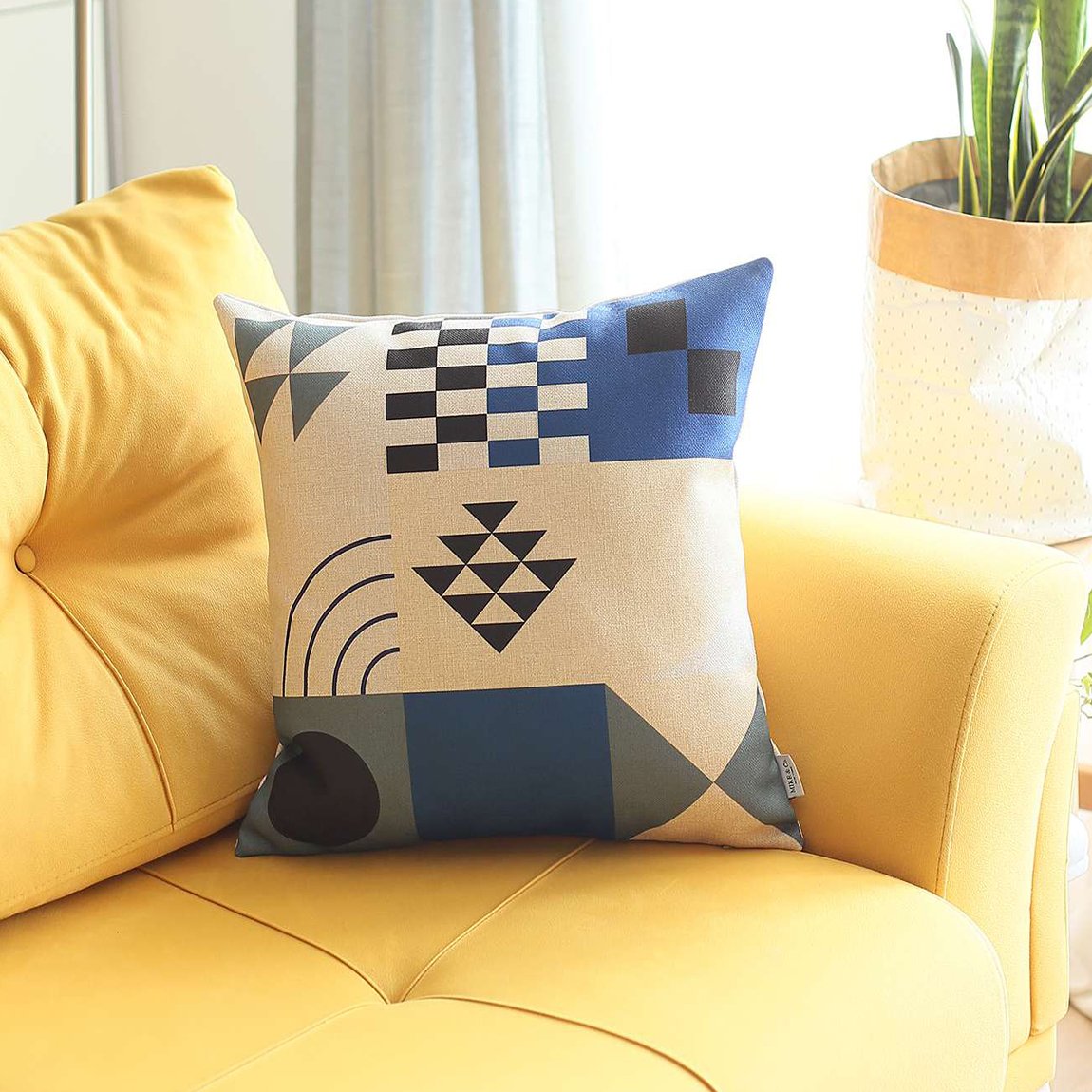 An 18" x 18" multi-color Bohemian Handmade Decorative Throw Pillow with an abstract jacquard pattern rests on a yellow sofa next to a potted plant.