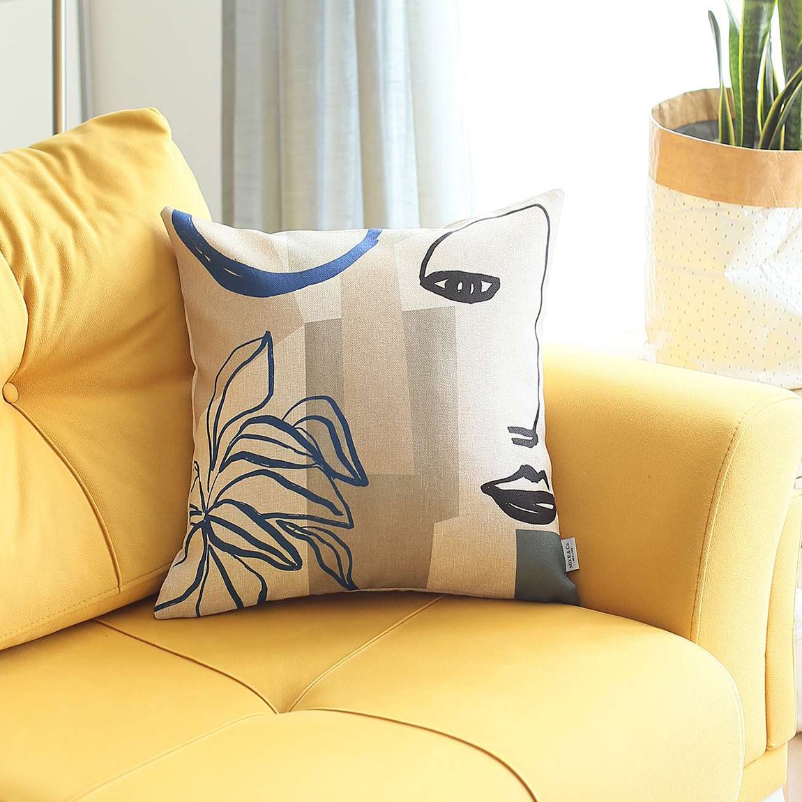 A yellow sofa is adorned with a Bohemian Handmade Decorative Throw Pillow, an 18" x 18" multi-color square featuring an abstract jacquard pattern. A lush plant graces the background, enhancing the cozy setting.