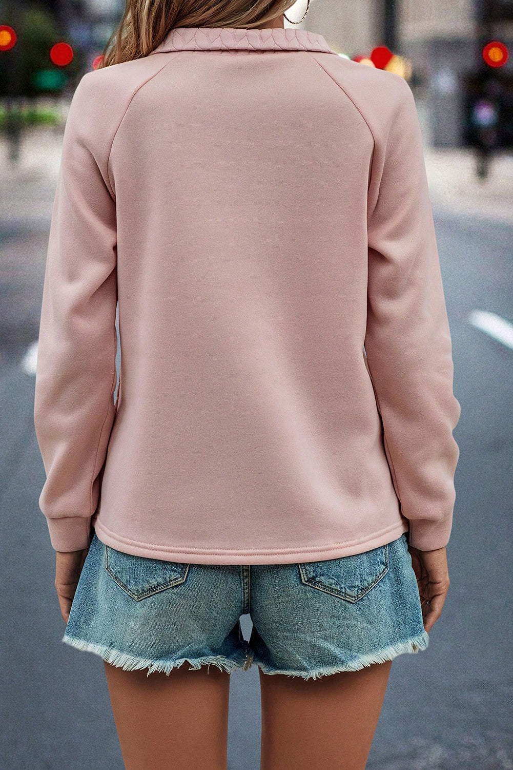 A person stands facing away from the camera, wearing a Pale Chestnut Textured Quarter Zip Raglan Sleeve Sweatshirt and blue jeans. The background features a grey textured wall and a tall pampas grass plant.