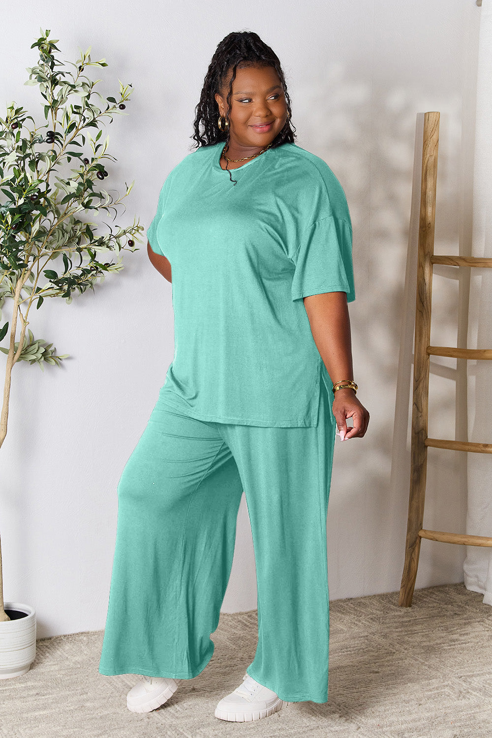 A woman stands near a potted plant and wooden ladder, wearing the Double Take Full Size Round Neck Slit Top and Pants Set in a bright pink two-piece outfit with matching wide-leg pants that are available in plus size and are easily machine washable.