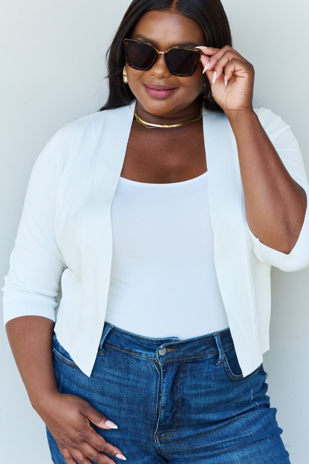 A stylish person wearing sunglasses, a Doublju My Favorite Full Size 3/4 Sleeve Cropped Cardigan in Ivory, and blue jeans poses in front of a plain background, effortlessly highlighting their versatile look.