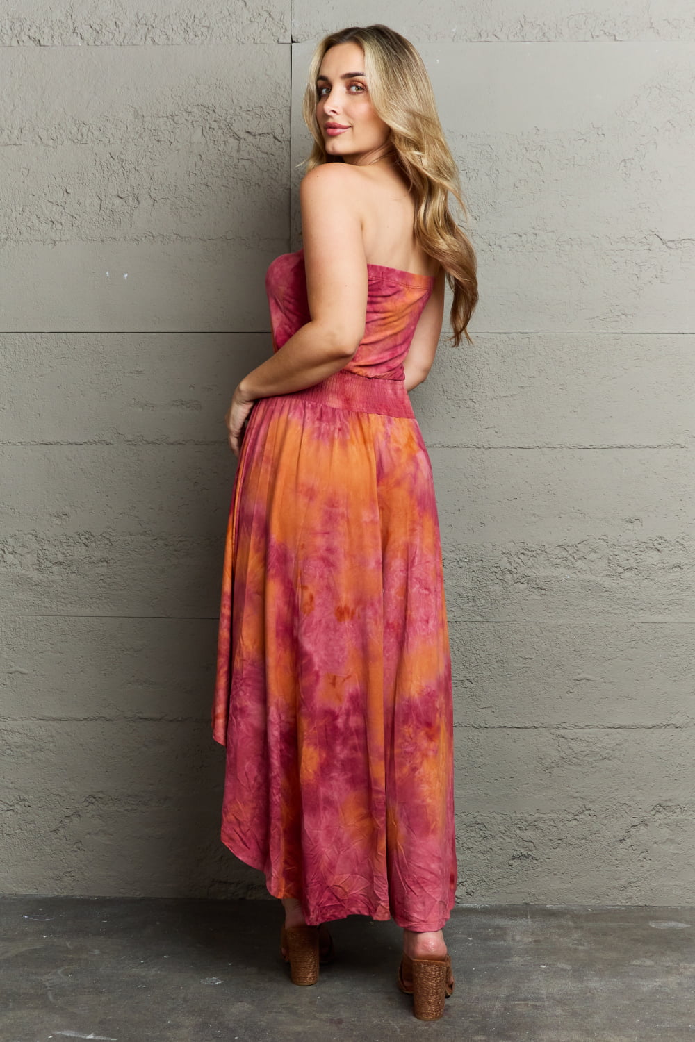 A woman with long blonde hair, wearing a Ninexis In The Mix Sleeveless High Low Tie Dye Dress in pink and orange and brown heels, stands against a gray textured wall, exuding bohemian charm.