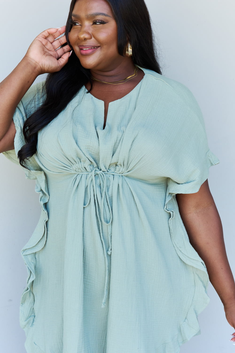 A woman is wearing the Ninexis Out Of Time Full Size Ruffle Hem Dress with Drawstring Waistband in Light Sage. Her long, dark hair falls gracefully as she smiles and looks to her left, standing against a plain background.