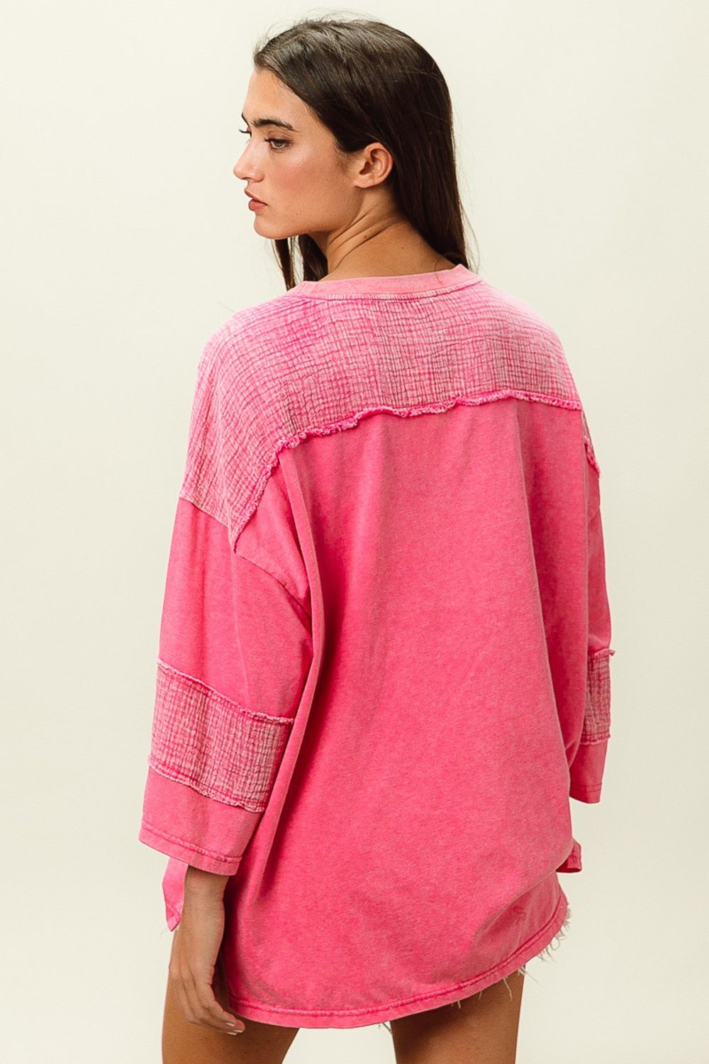 A young woman with long dark hair wears a bright pink BiBi High-Low Washed T-Shirt featuring a front pocket, paired with white frayed denim shorts. She poses against a plain background, effortlessly combining comfort and style in her trendy wardrobe.