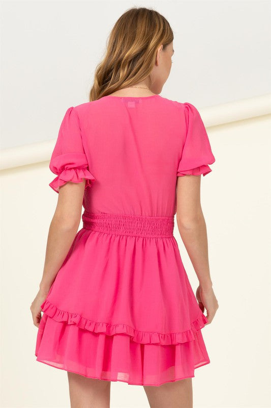 A woman is wearing the "Days Like This Tie-Front Mini Dress" in a vibrant pink color, featuring short ruffled sleeves, a prominent bow at the chest, and a tiered skirt with a smocked waist.