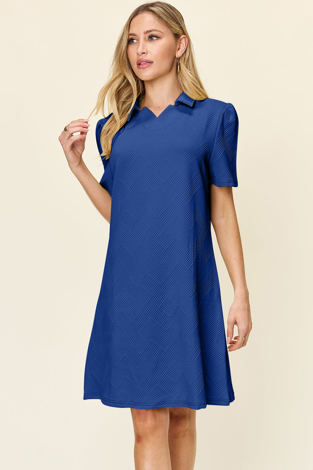 A woman wearing the Double Take Full Size Texture Collared Neck Short Sleeve Dress, featuring a geometric pattern in purple and made from soft polyester, stands against a neutral background. She has long, light hair and is looking slightly to the side. The dress is available in sizes S-3XL and is easy to care for—just machine wash cold.