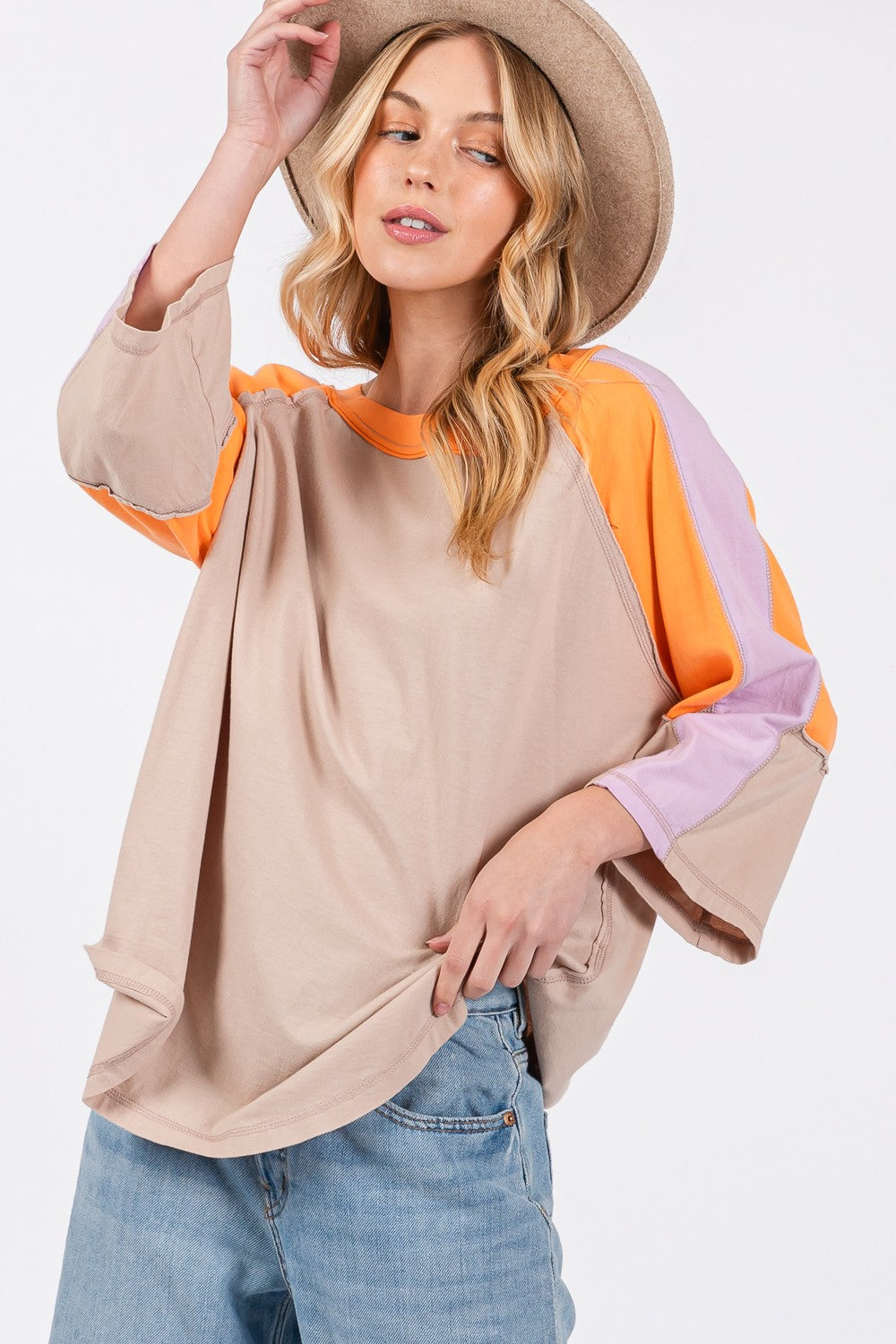 Person wearing a chic modern look with the SAGE + FIG Color Block Curved Hem T-Shirt in beige, featuring orange and purple accents, paired with blue jeans and a hat, poses against a plain background.