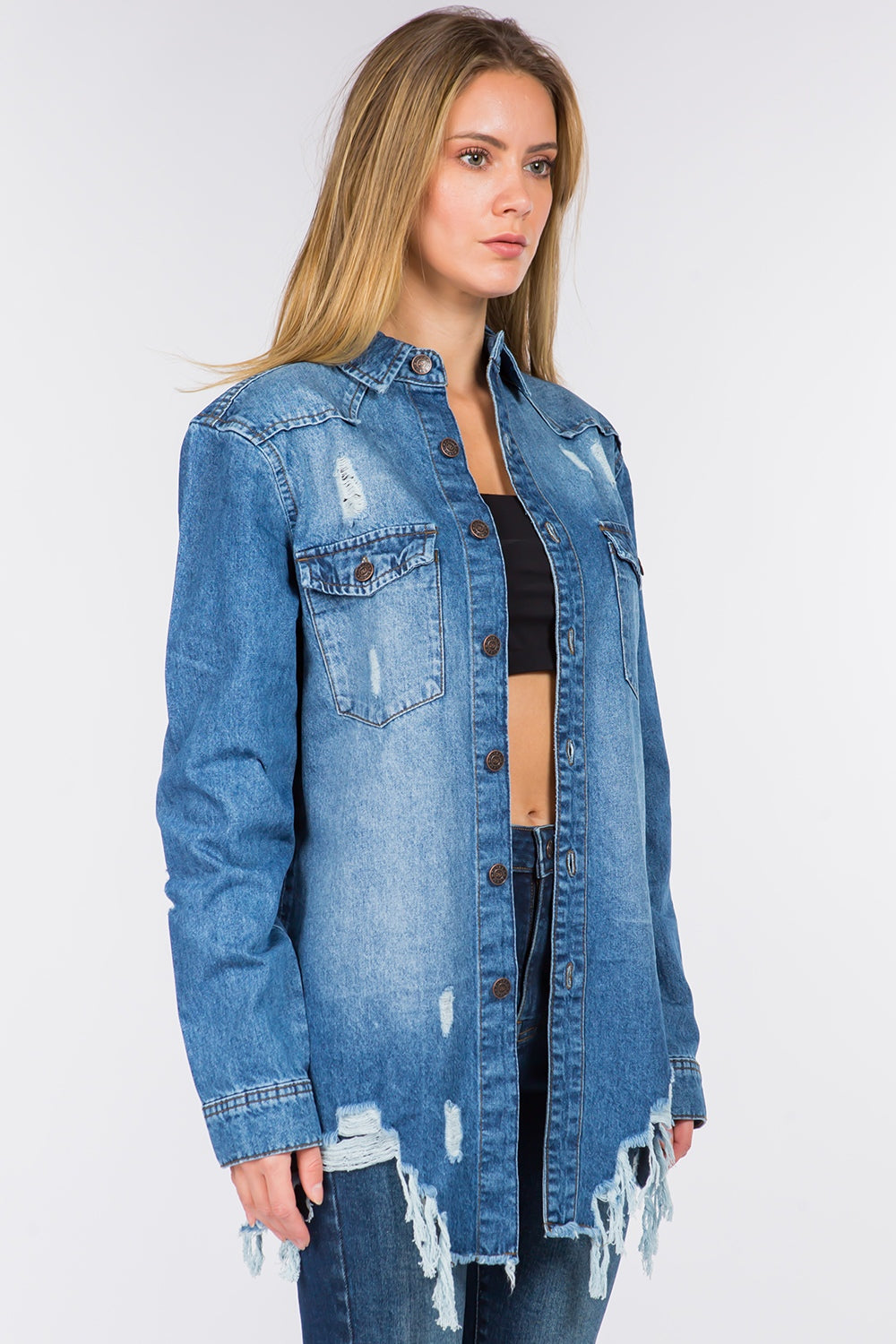 A person with long hair is standing, wearing an American Bazi Distressed Frayed Hem Denim Jacket over a black top and denim jeans with a frayed hem.