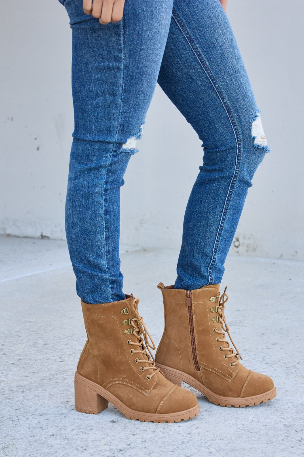 A person wearing distressed blue jeans and Forever Link Lace-Up Zipper Detail Block Heel Boots stands on a concrete surface.