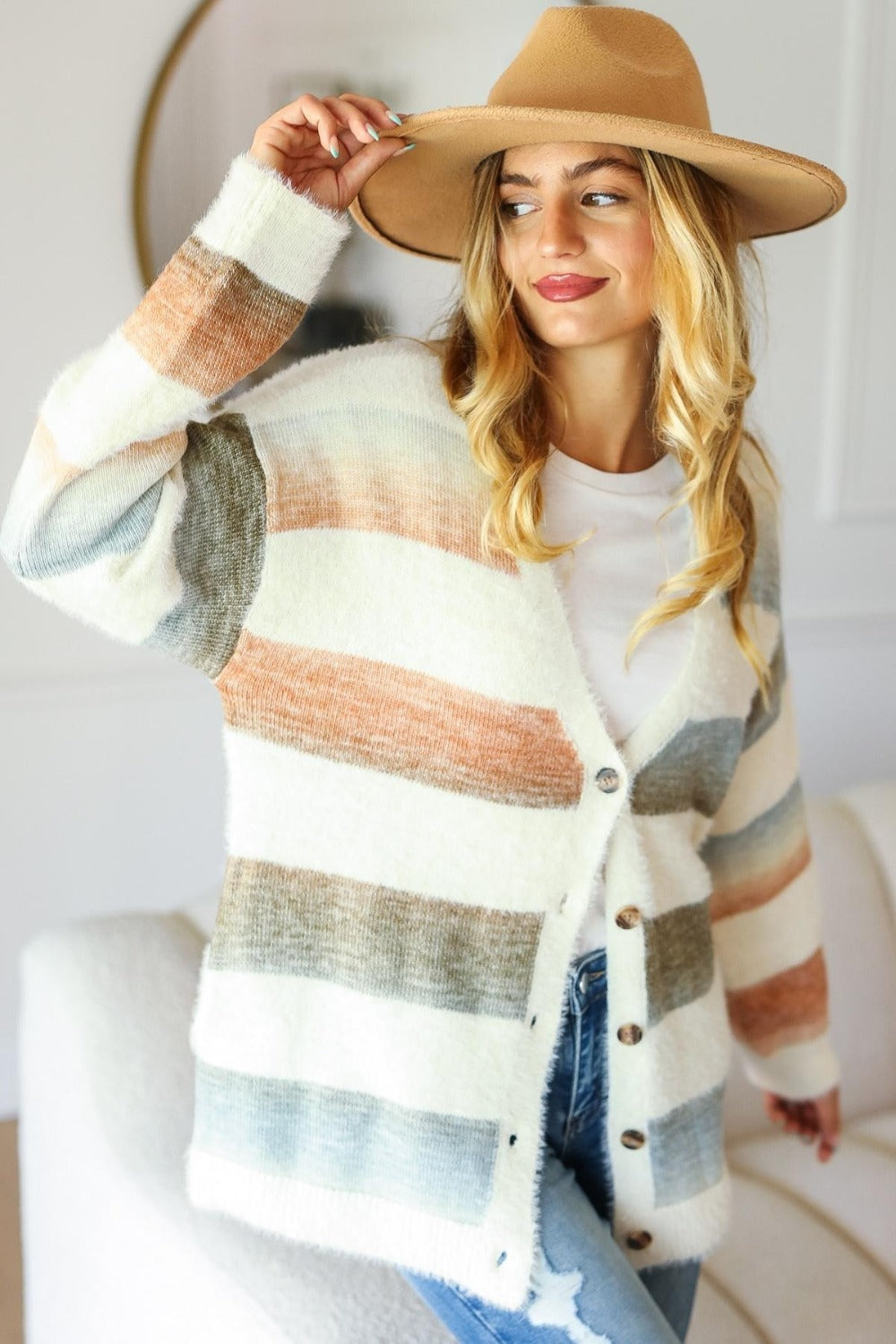 A woman with long blonde hair, wearing a wide-brimmed hat, Haptics Full Size Button Down Stripe Soft Fuzzy Sweater Cardigan, and jeans stands indoors, tilting her hat with one hand and smiling.