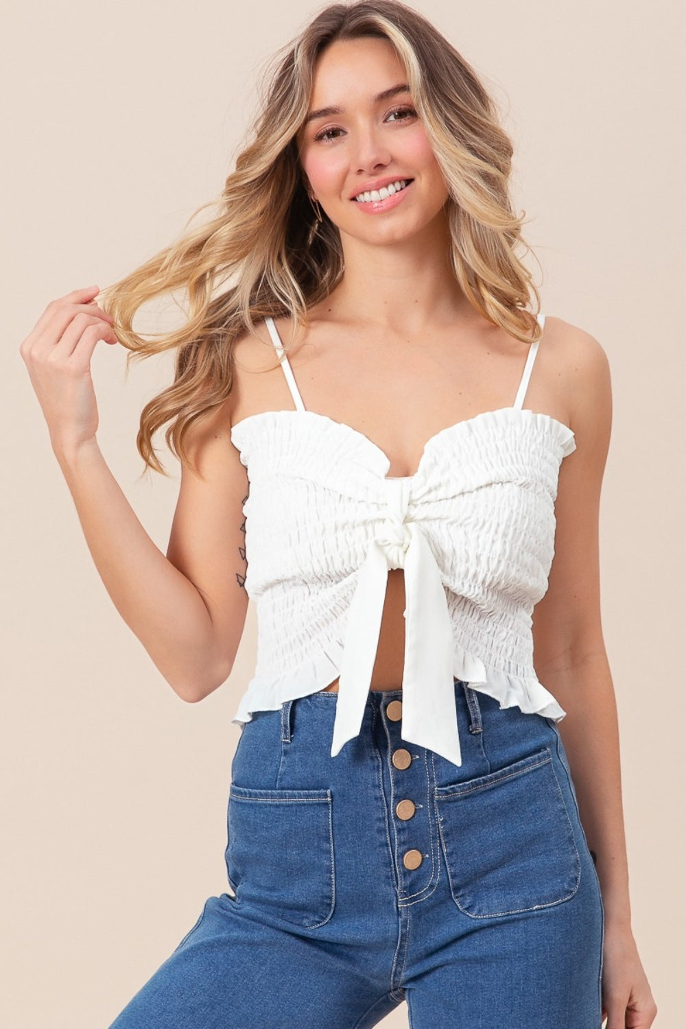 A woman wearing the BiBi Ruffled Smocked Ribbon Detail Cami in white, paired with blue high-waisted jeans, is posing against a neutral background.